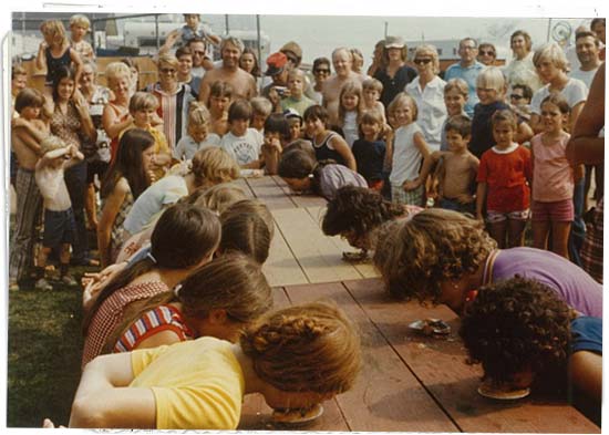 People watch youth, seated at picnic tables, particpate in a 1960s pie-eating contest.